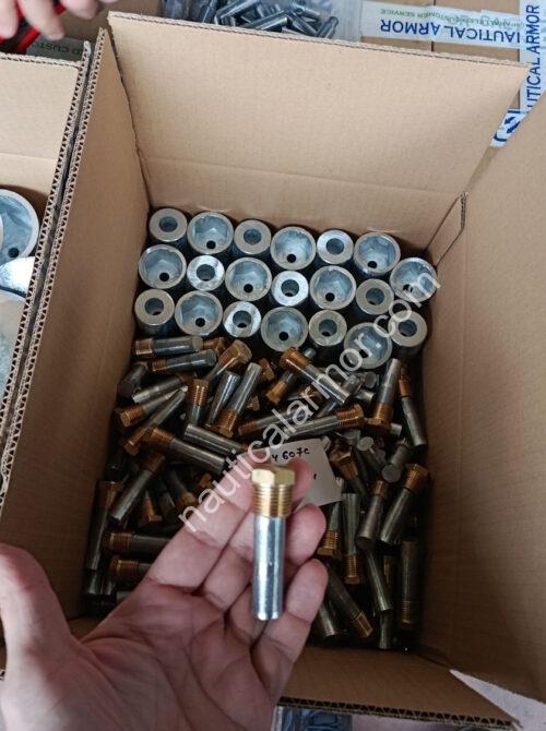 Nautical Armor hand holding a bolt above boxed sacrificial anodes and hardware with Nautical Armor branded tape visible.