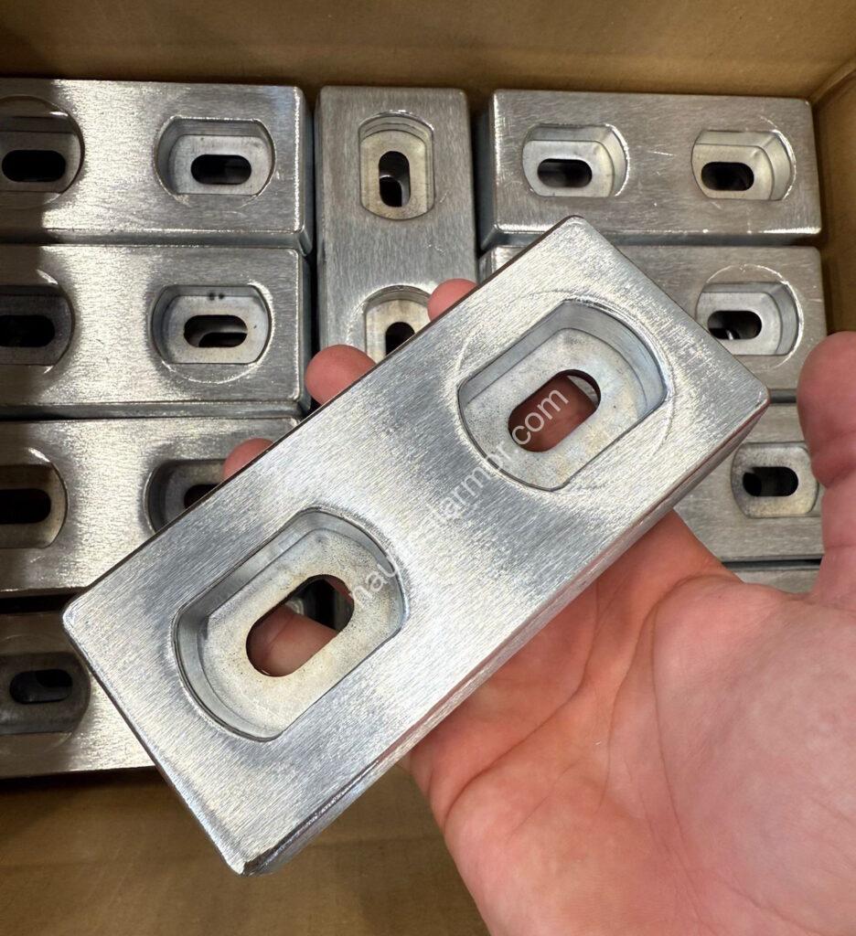 Hand holding a rectangular Nautical Amor bolt-on zinc BLT-800267 anode above a box filled with the same anode model.