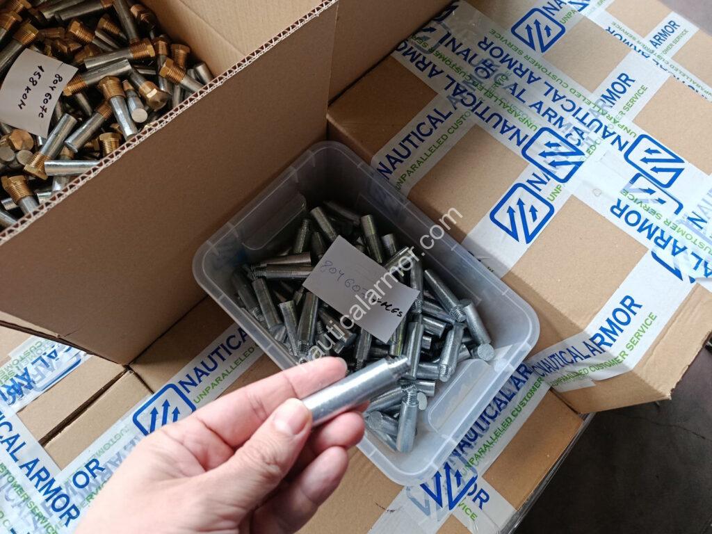 Nautical Armor hand holding a pencil engine anode with boxes and labeled bins of anodes and fasteners.